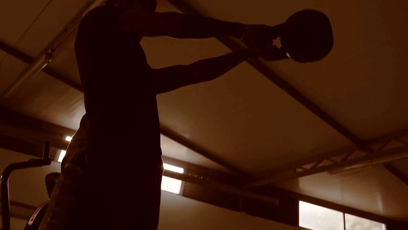 A member at the top of a kettlebell swing, lit from behind against the roof of the gym.