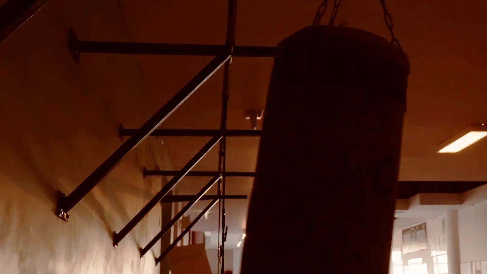 A heavy bag hanging on its chains in the corner of the gym, wall rails running away into the dark.